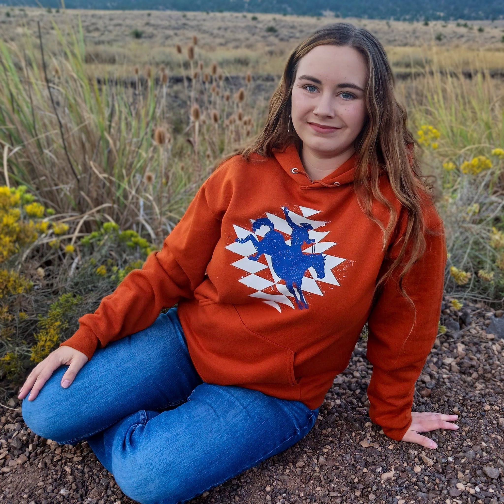 Cowgirl wearing an orange hoodie with a graphic design of a cowboy, sitting in a a field with a ranch landscape behind her.