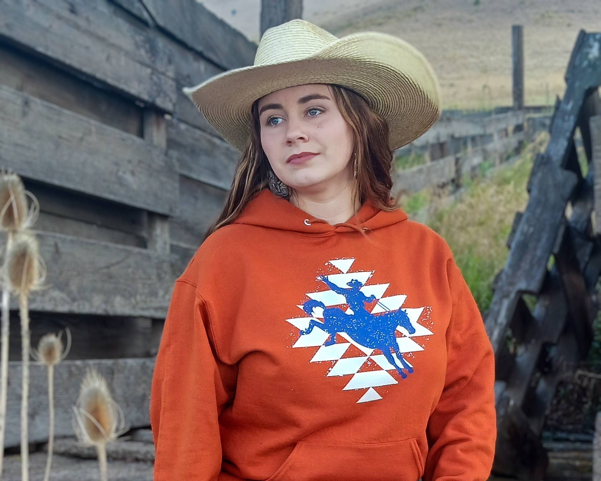 Cowgirl sitting on a rustic fence, wearing an orange hoodie with a graphic design of a cowboy riding a horse on top of an Aztec print.