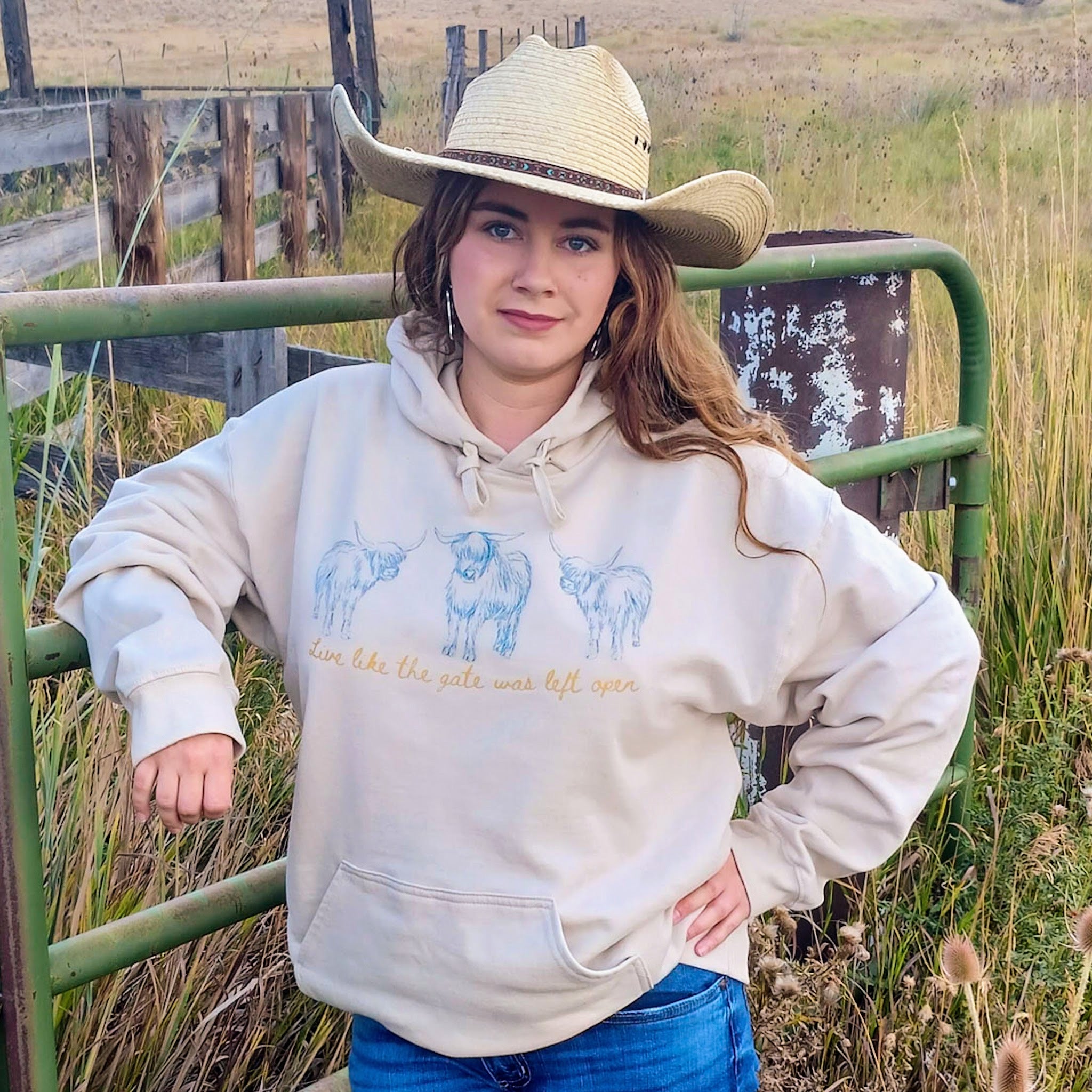 Cowgirl wearing a beige hoodie with blue highland cows and yellow text that says “Live like the gate was left open” design while standing by a gate in a field.