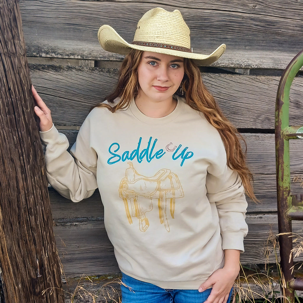 Cowgirl wearing a beige sweatshirt with 'Saddle Up' text and a yellow vintage saddle graphic, standing against a worn wooden cattle chute background.