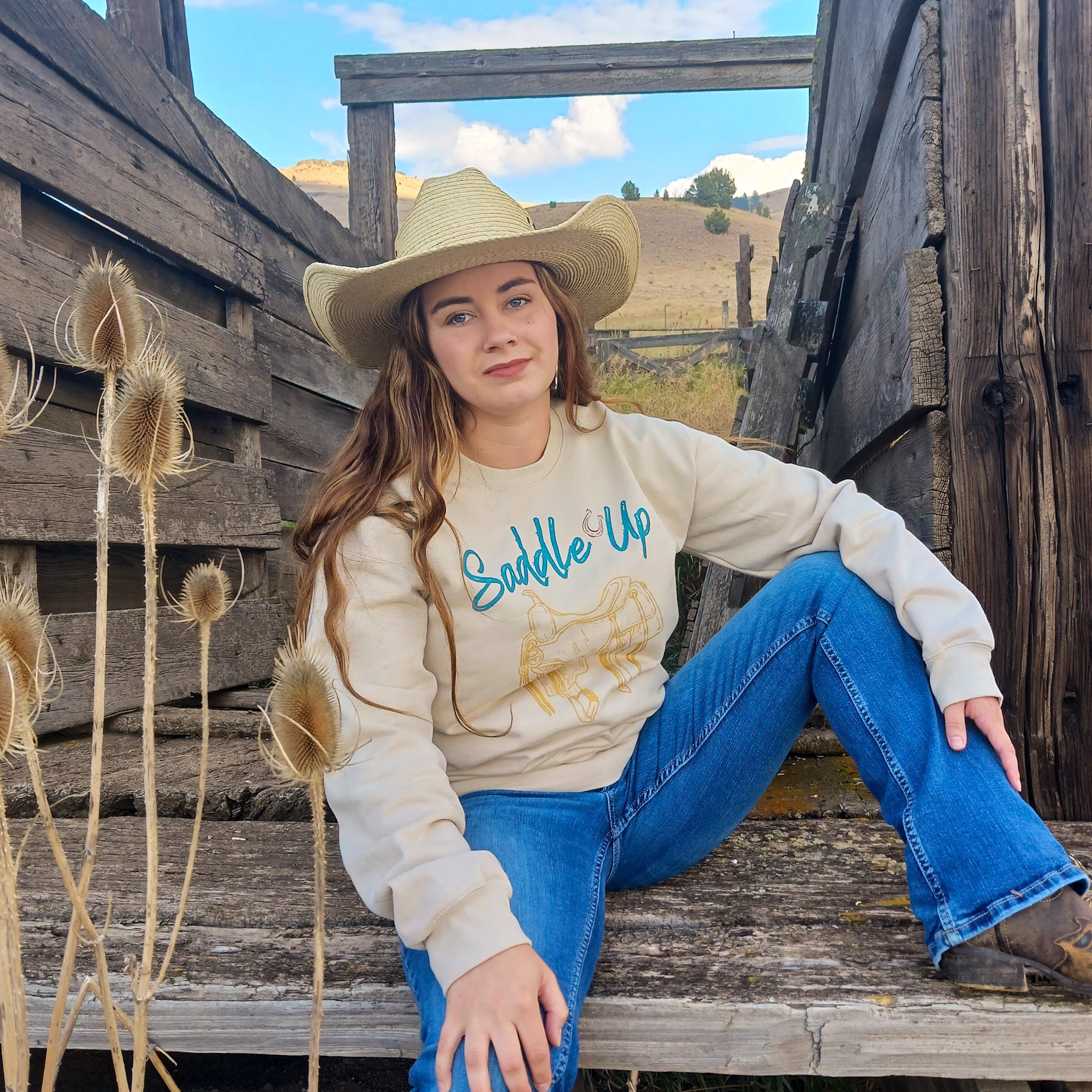 A cowgirl wearing a 'Saddle Up' sweatshirt sitting on wooden cattle chute with a rustic ranch. 