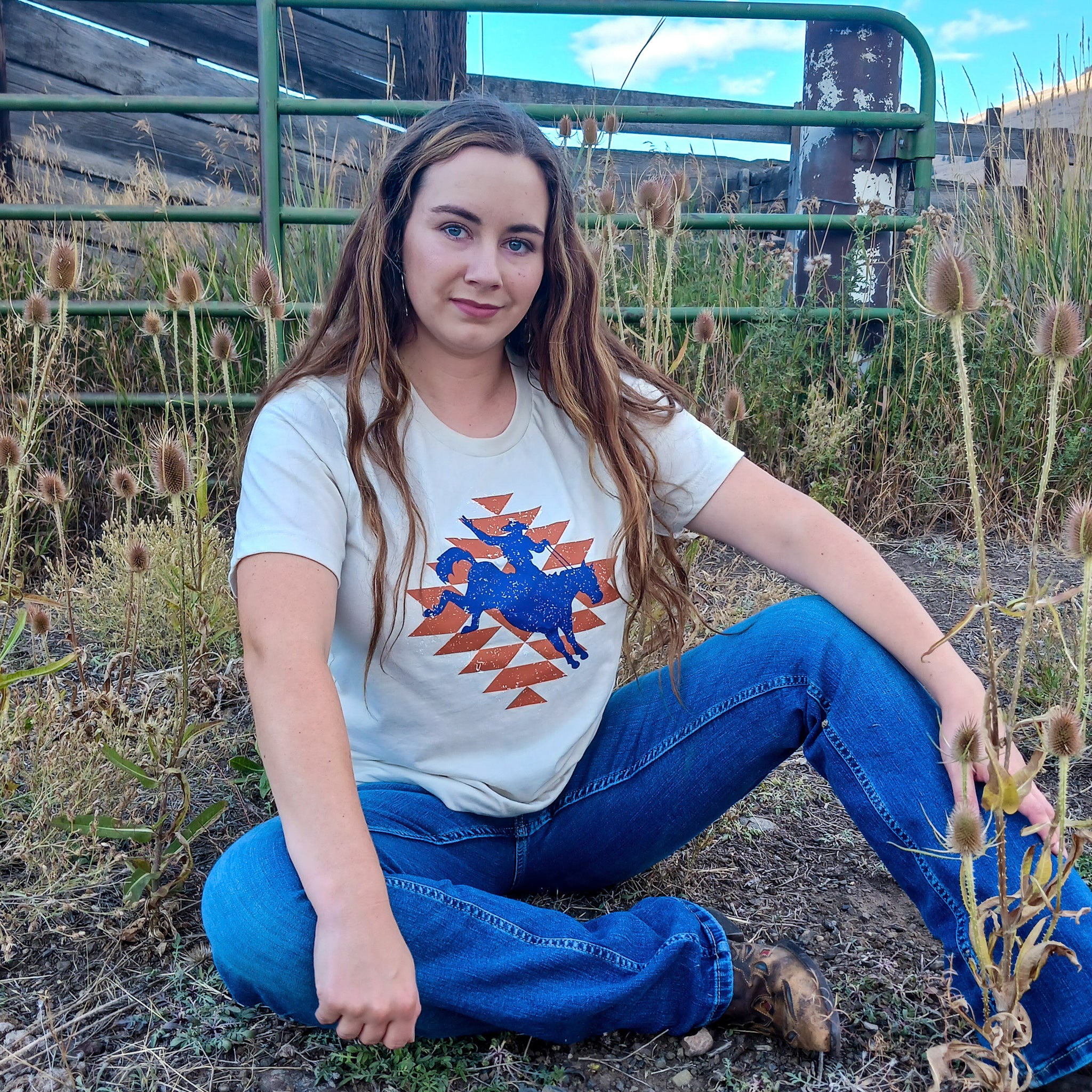 Woman sitting on the ground wearing a t-shirt with a Western Aztec cowboy graphic design in an outdoor ranch setting.