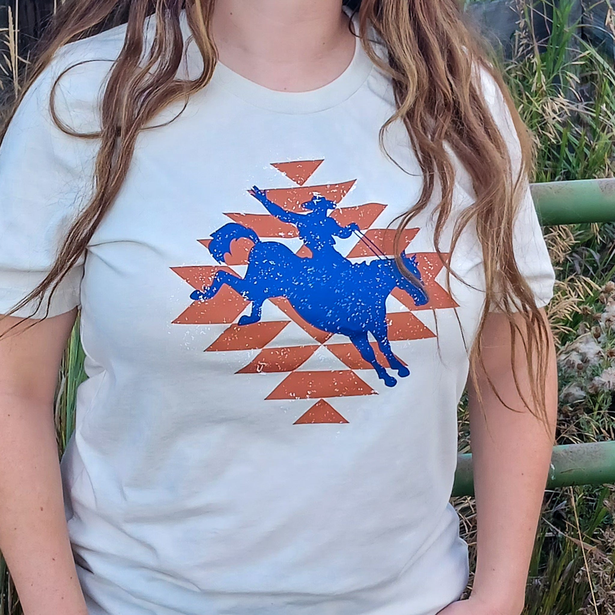 Women wearing an off white t-shirt with a blue graphic of a person on a horse on top of an orange geometric aztec patterns. 