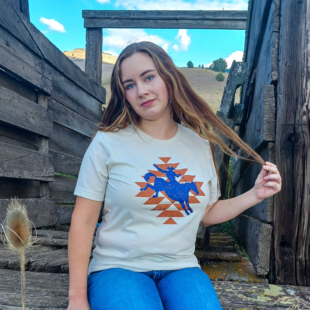 A cowgirl wearing a t-shirt with a graphic design of a horse and cowboy in blue on top of an orange Aztec geometric print, standing in front of wooden structures cattle chute that is made of weathered wood with a ranch in the background.