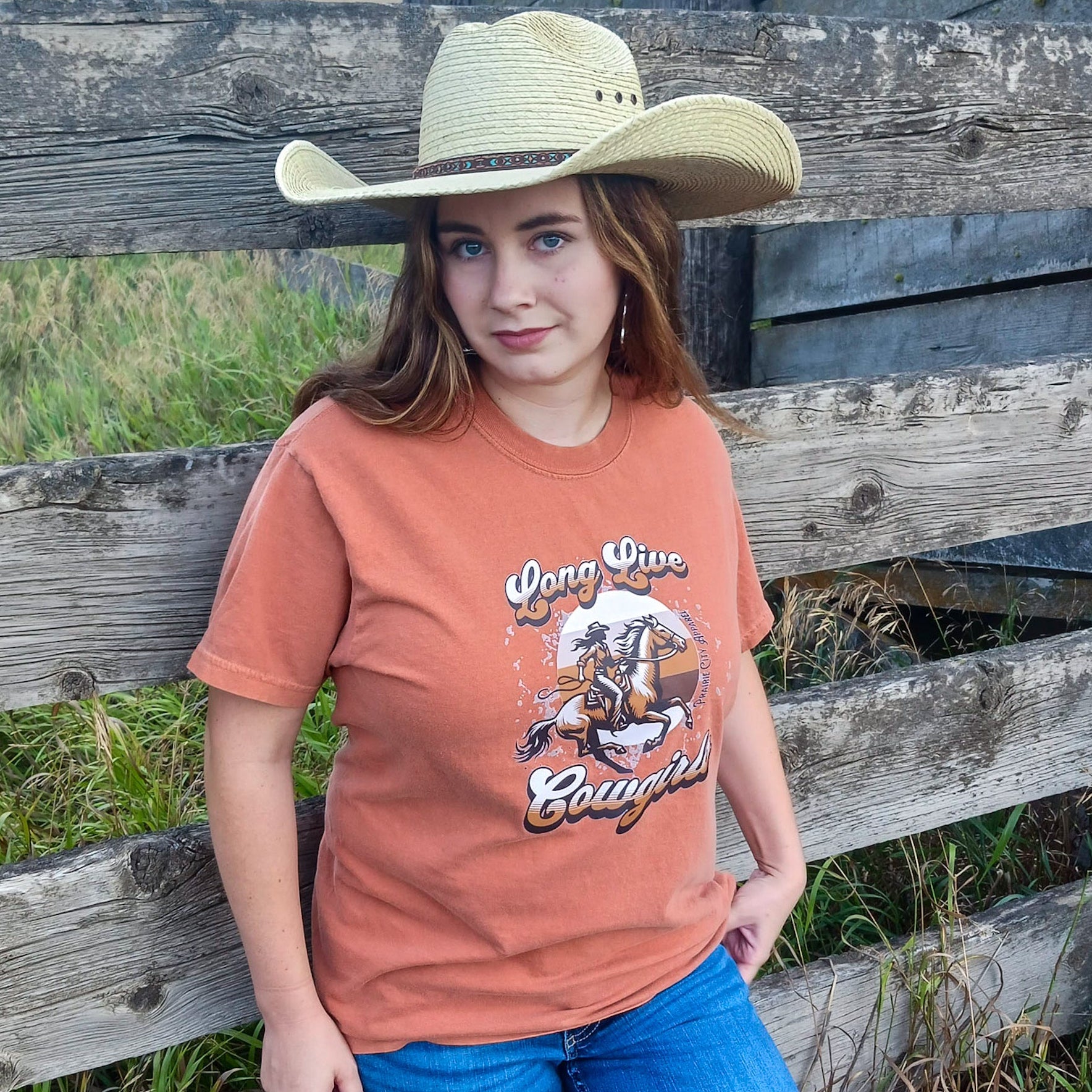 Cowgirl wearing a dark orange Western Graphic tee, with a brown cowgirl on a horse graphic with the words “long live cowgirls” written on it.