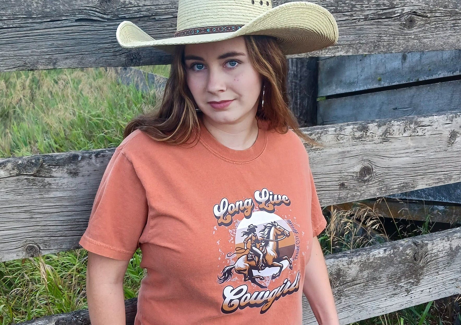 Cowgirl wearing a dark orange Western Graphic tee, with a brown cowgirl on a horse graphic with the words “long live cowgirls” written on it.