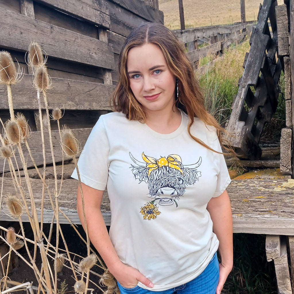 Cowgirl wearing an off white t-shirt with a yellow whimsical highland cow graphic design, with a bow and a sunflower, standing in front of a wooden cattle chute with a ranch in the background.