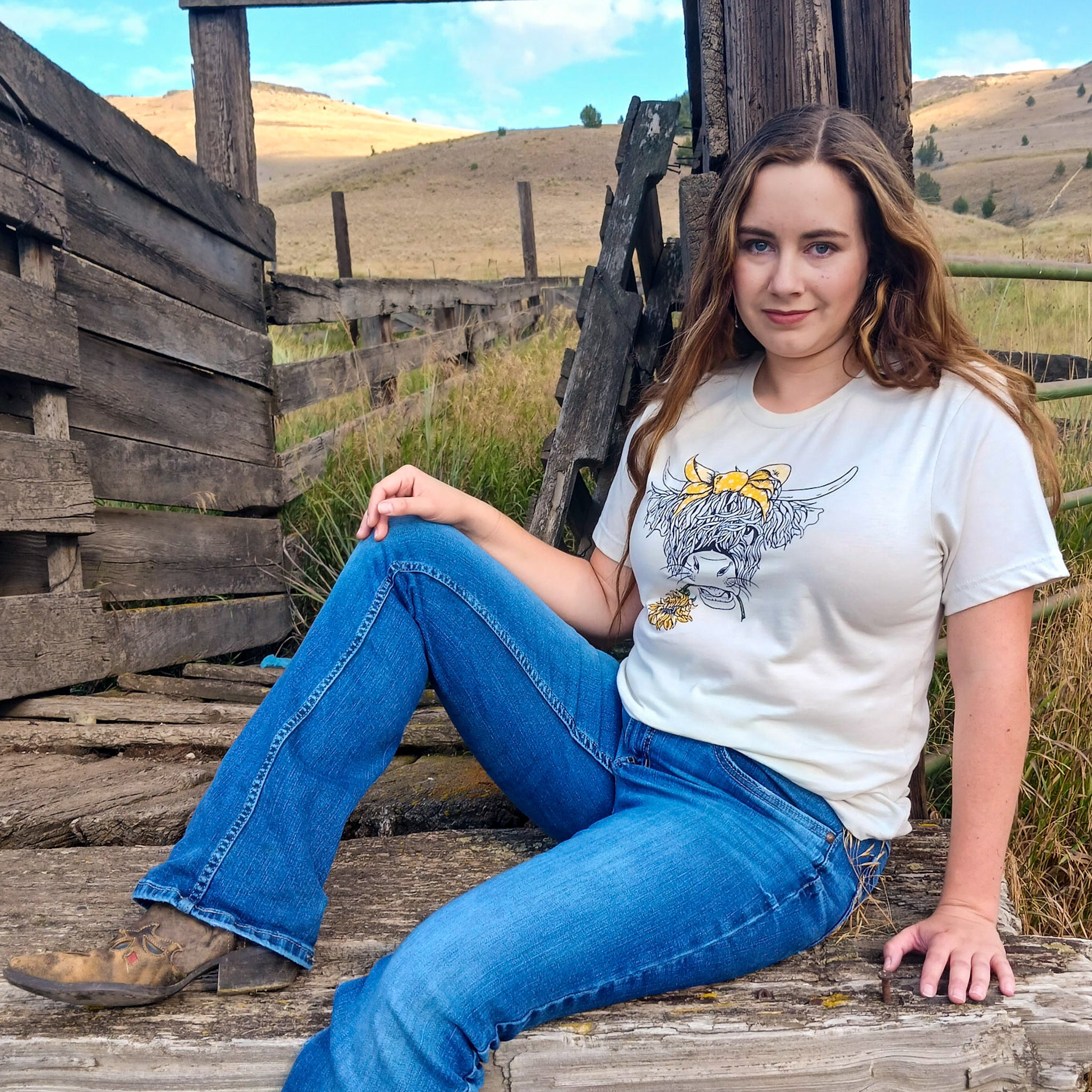 Cowgirl sitting on wooden cattle chute wearing a Wester Highland Cow tee with a sunflower