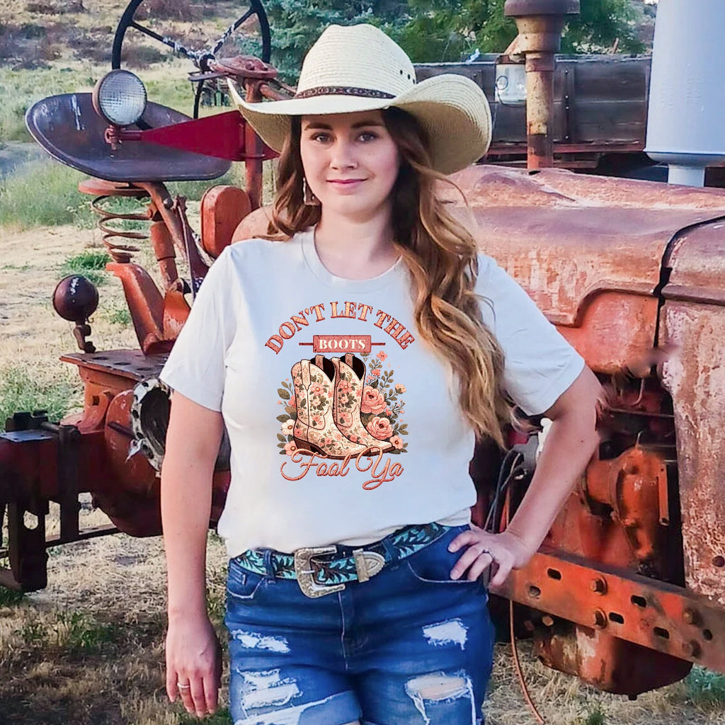 Women wearing a t-shirt with a graphic that says "Don't Let The Boots fool Ya!" With xowgirl boots and pink roses, while standing in front of an old tractor.