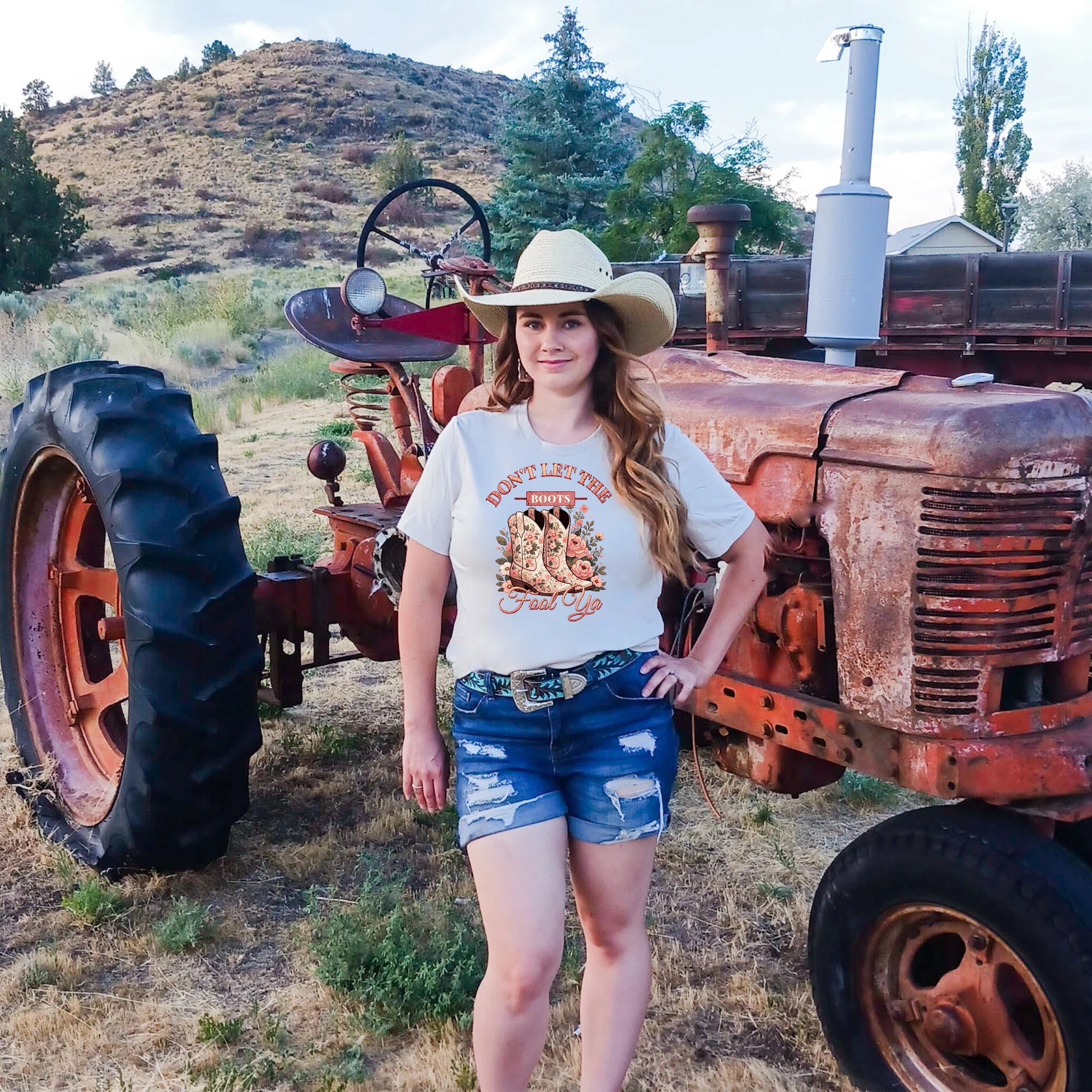 Woman in a cowboy hat and graphic t-shirt standing next to an old tractor in a rural setting.