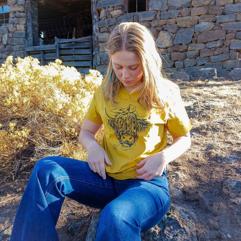A cowgirl wearing a yellow Western graphic tee with a highland cow with a daisy in it mouth on the tee with a rustic barn in the background