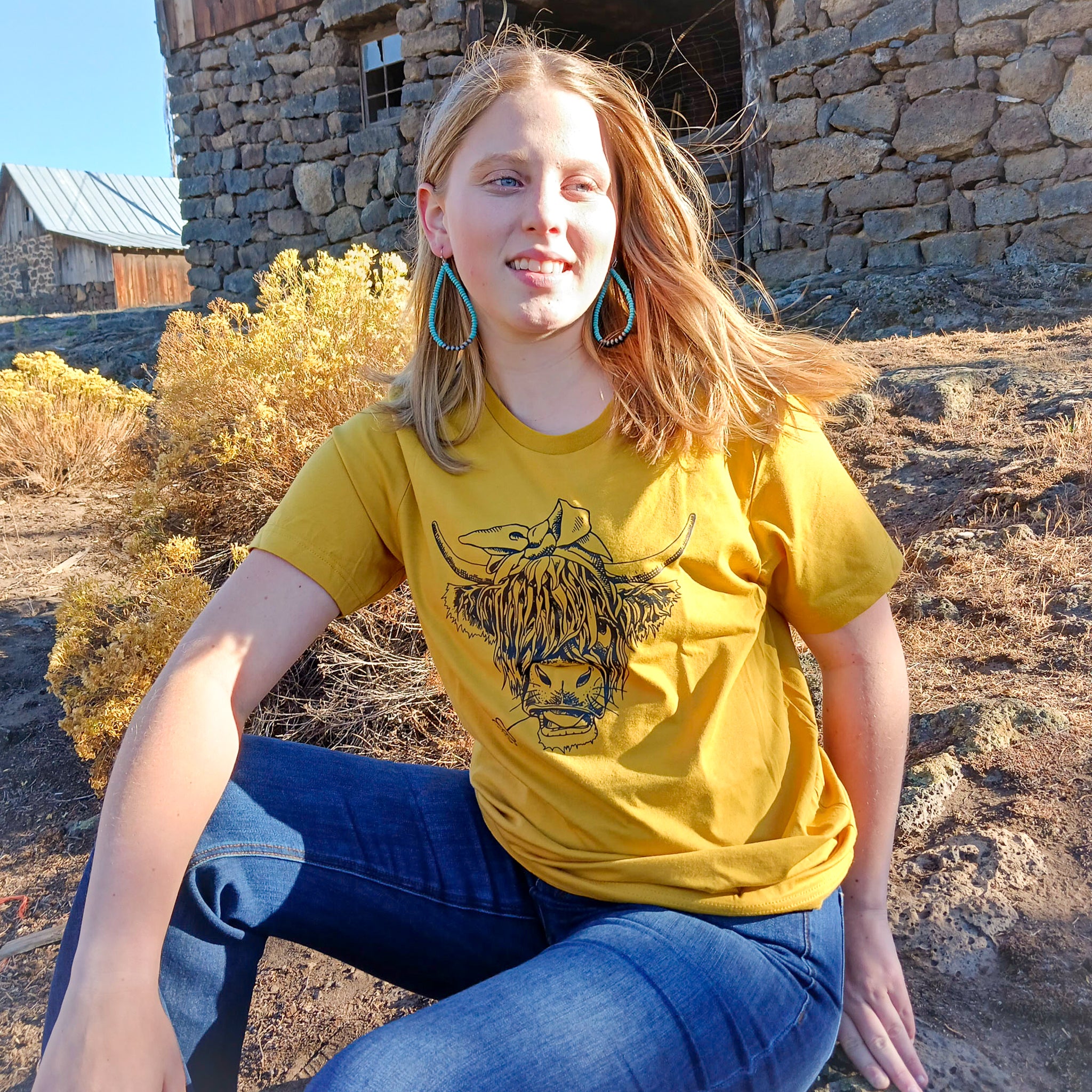 A cowgirl wearing a yellow Western graphic tee with a highland cow with a daisy in it mouth on the tee with a rustic barn in the background