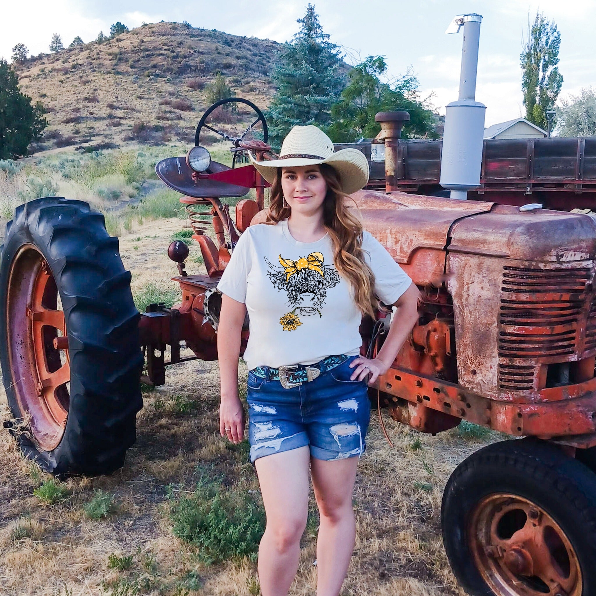 Woman standing in front of an old tractor with a mountainous landscape in the background wearing a Highland cow western graphic tee