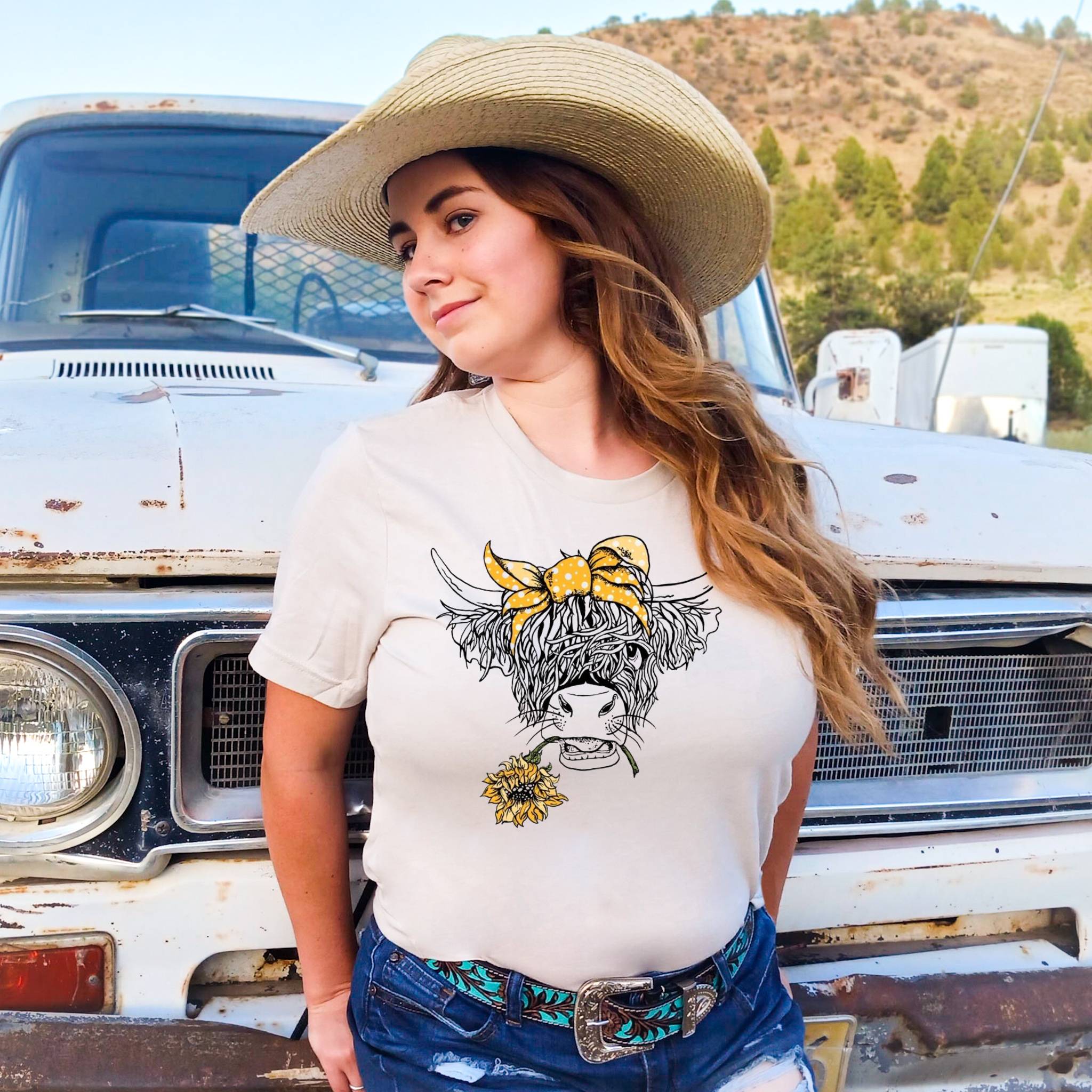 Woman wearing a off white t-shirt with a highland cow graphic in front of an old truck.