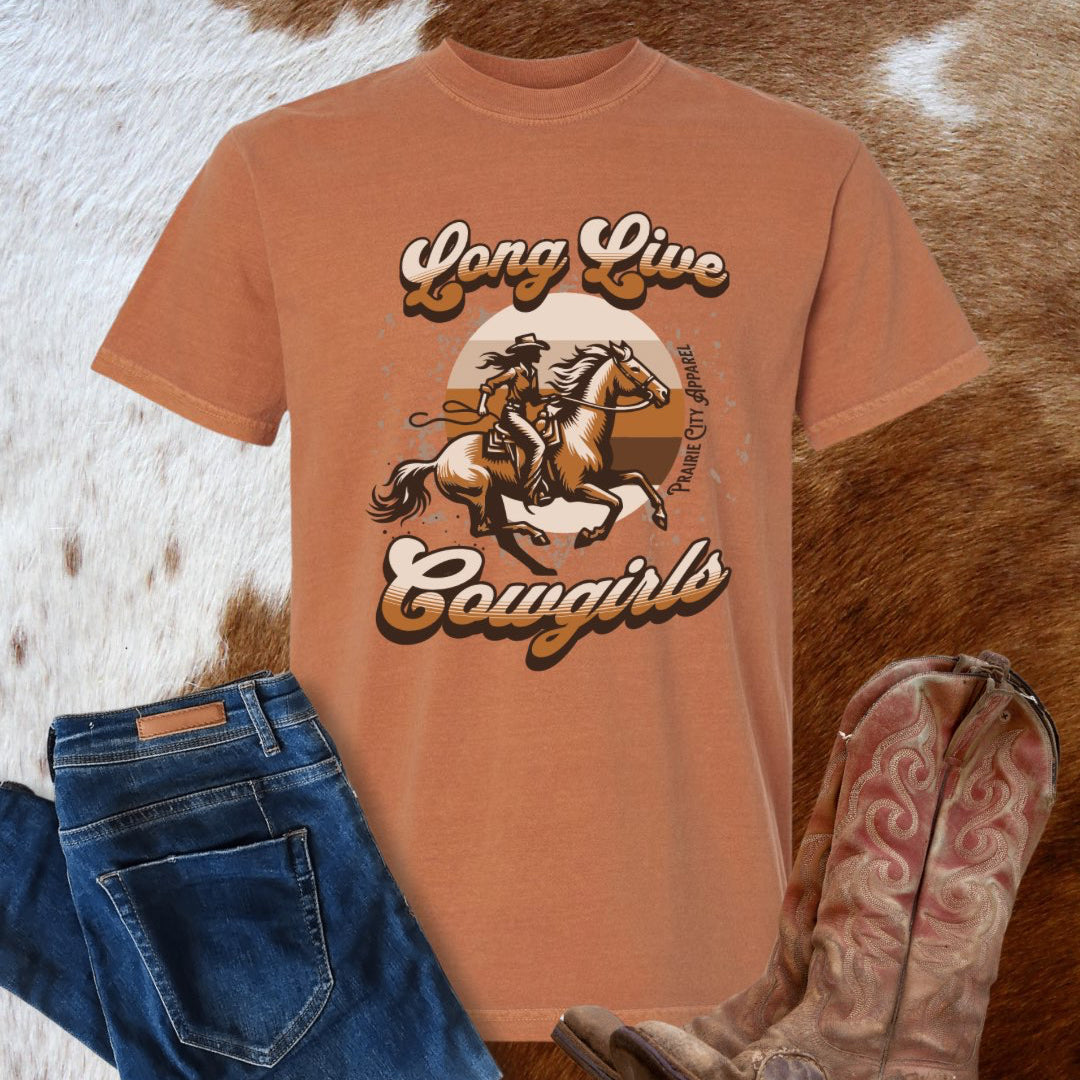 Orange t-shirt with 'Long Live Cowgirls' graphic, blue jeans, and cowboy boots on a textured surface.