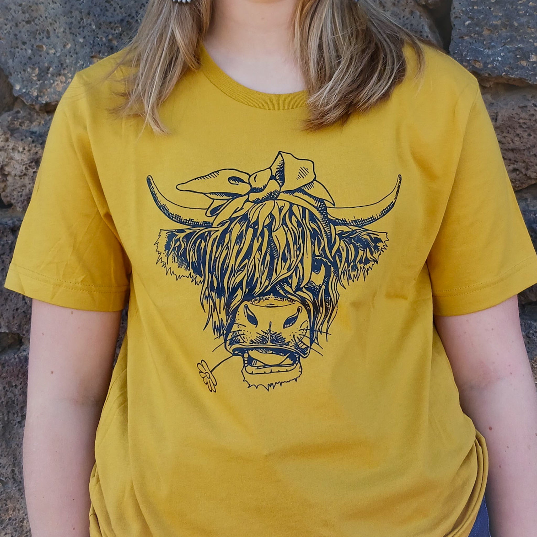 A cowgirl wearing a yellow Western graphic tee with a highland cow with a daisy in it mouth on the tee