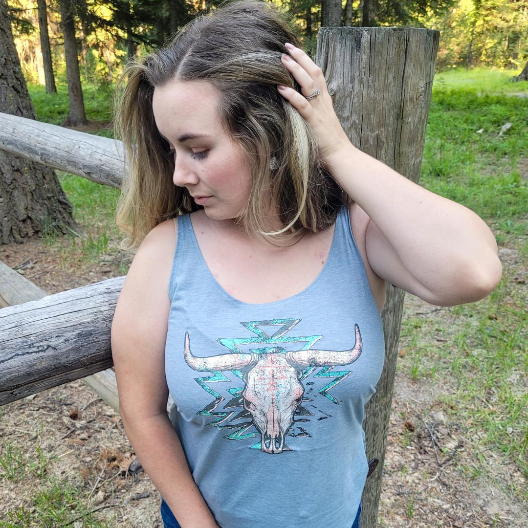 Cowgirl wearing a light blue western tank top with an Aztec bull skull graphic with a fence behind her
