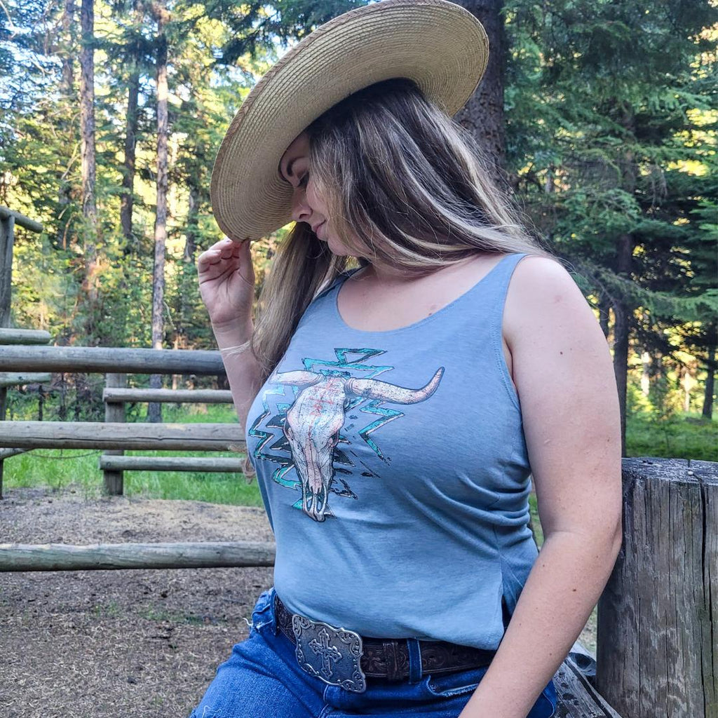 Cowgirl posing in a cowgirl hat on a fence by a horse pen with a light blue western graphic tank top with a Aztec bull skull design on it.